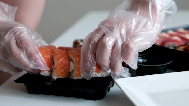 Female hands in transparent disposable gloves carefully transfer Philadelphia sushi rolls from black delivery box onto a white plate. scene captures neat food presentation, home dining aesthetics. 