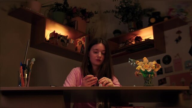 Young caucasian focused woman sits at the table arranges blossoms with dedication in cozy studio at evening. Assembling flowers from a construction set. Hobbies for adults, fine motor skills
