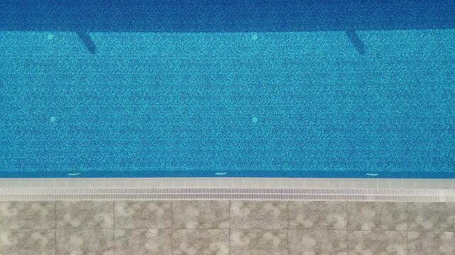 Top down aerial view moving along a beautiful empty blue water swimming pool with shimmering light reflections in a serene hotel resort or private villa environment