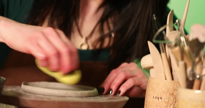 A close-up of a potter's hands using a damp yellow sponge to smooth and refine the edges of a raw clay disk on a rotating pottery wheel.

