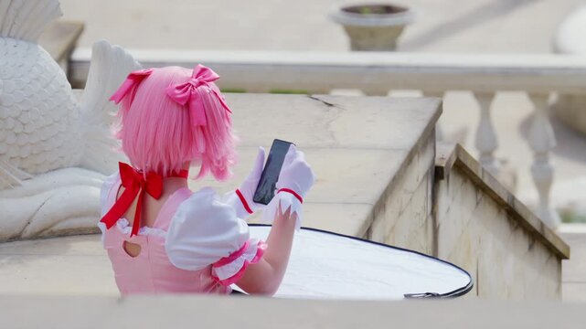Back view of a young woman in pink cosplay costume holding a smartphone near a reflector in an outdoor location