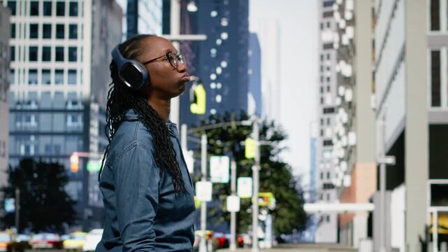 Cheerful black woman performing stylish breakdance moves with headphones, dancing around on sidewalk in town square. Jolly girl having fun with groovy moves, trendy vibrant routine downtown.