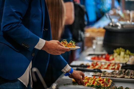 Man in blue blazer holding plate with appetizers at a buffet table featuring various dishes and a blurred background of people and food items