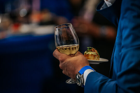 Man in blue suit holding a glass of white wine and a plate with a small appetizer at a social gathering with blurred background elements