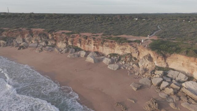 Aerial view of the Calas de Roche cliffs at sunset in Conil, Spain. Cinematic drone shot of the red sandstone cliffs and turquoise Atlantic Ocean at golden hour.