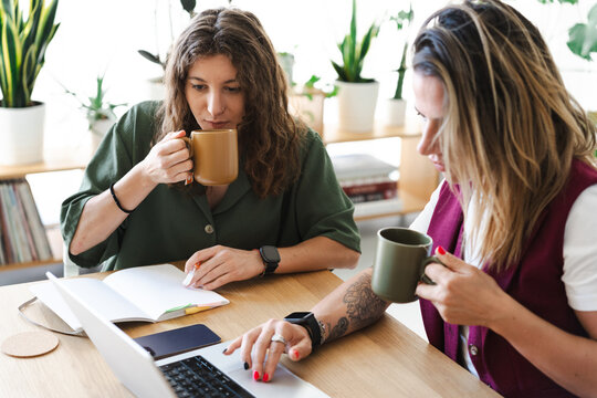 Two women are working together at a table, one is typing on a laptop and the other is drinking from a mug.