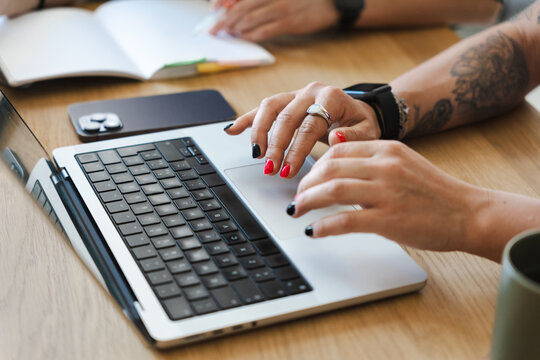 Hands with painted nails use a laptop and a smartphone on a wooden desk, with an open notebook and a mug nearby.
