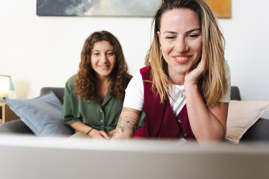 Two women are sitting on a couch and looking at a laptop. One woman is in the foreground, smiling and resting her chin on her hand. The other woman is in the background, also smiling.