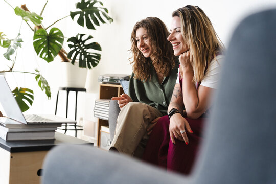 Two women are sitting on a sofa, smiling and looking at a laptop. One woman is pointing at the screen.