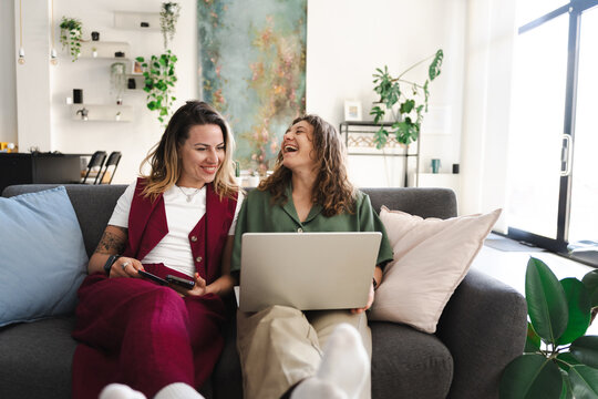 Two women laugh while sitting on a sofa, one holding a phone and the other a laptop, in a bright, modern living room with plants.
