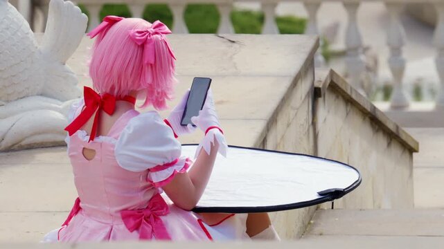 Back view of a young woman in pink cosplay costume holding a smartphone near a reflector in an outdoor location