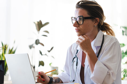 A female doctor in glasses and a white coat sits at a desk, holding a pencil and looking thoughtfully at a laptop screen.