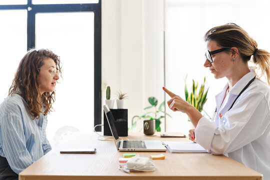 A doctor in a white coat consults with a patient, pointing to a notebook on the desk.