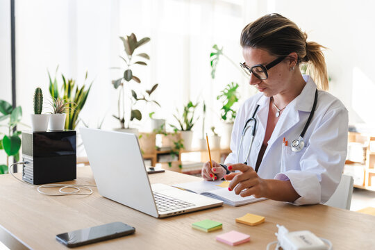 A female doctor in a white coat with a stethoscope around her neck is writing in a notebook at her desk, with a laptop and plants in the background.