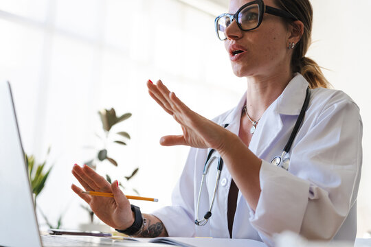 A female doctor in a white coat and stethoscope gestures while speaking to a laptop, holding a pencil and looking engaged in a virtual consultation.