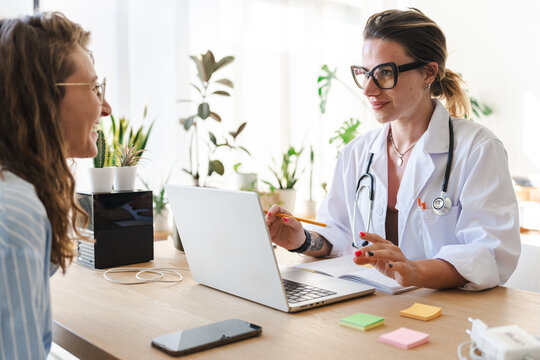 A doctor in a white coat consults with a patient, using a laptop and notes to discuss medical information in a bright, modern office setting.