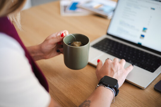 A woman's hands are shown as she works on her laptop and drinks tea. She wears a smartwatch and jewelry.