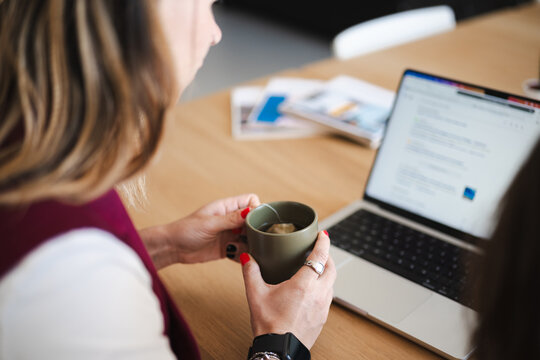A woman holds a cup of tea while working on her laptop at a wooden desk, surrounded by papers and books.