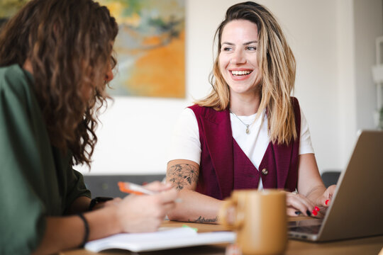 Two women collaborate at a table, one typing on a laptop while the other takes notes, sharing ideas and working together on a project.