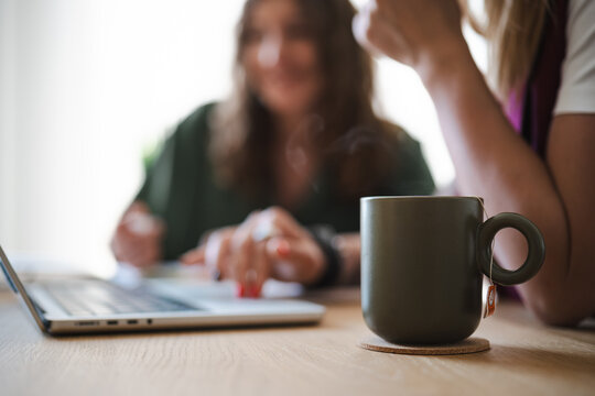 Two women collaborate on a project at a table, one typing on a laptop while the other holds a cup of tea.