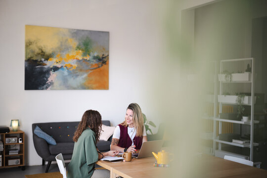 Two women collaborate at a table, one using a laptop while the other writes in a notebook, in a bright, modern office space.