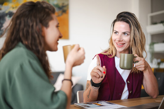 Two women are having a conversation over coffee at a table, one gesturing with her hand while holding a mug.