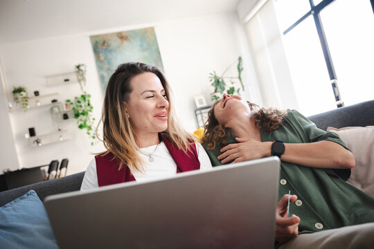 Two women are relaxing on a sofa, one looking at a laptop while the other leans back with her eyes closed, sharing a moment of connection.