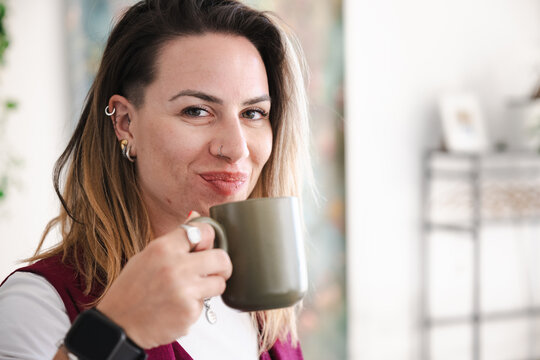A woman with piercings and a smartwatch smiles as she holds a mug, enjoying a moment of relaxation indoors.