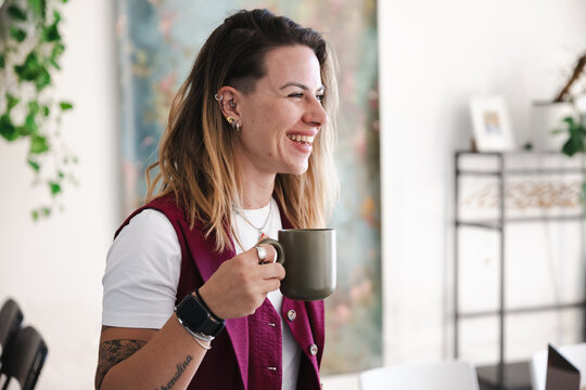 A woman with tattoos and piercings smiles while holding a mug, enjoying a moment of relaxation in a bright, modern space.