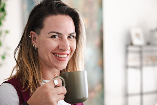 A smiling woman with piercings and a nose ring holds a mug, enjoying a warm beverage indoors.