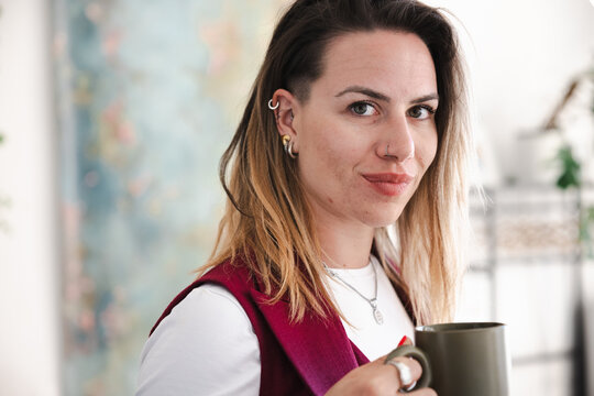 A woman with piercings and styled hair holds a mug, looking towards the camera with a relaxed expression.