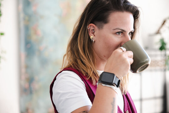 A woman with multiple ear piercings and a smartwatch drinks from a mug, looking thoughtfully into the distance.