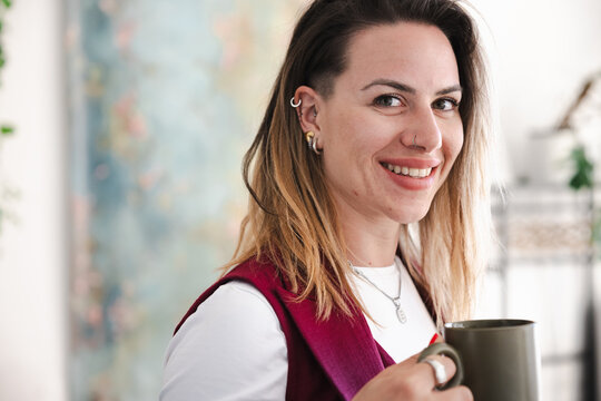 A smiling woman with piercings and styled hair holds a mug, looking towards the camera with a relaxed expression.