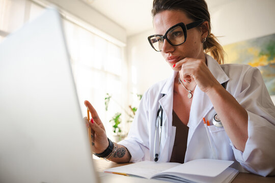 A focused female doctor in a white coat reviews information on a laptop, holding a pencil and resting her chin on her hand.