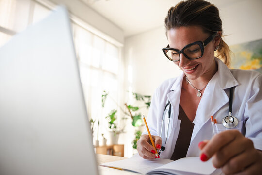 A female doctor wearing glasses and a white coat with a stethoscope around her neck writes in a notebook with a pencil.