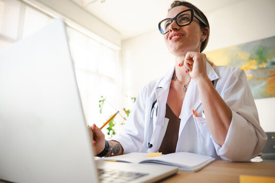 A female doctor in a white coat and glasses is working at her desk with a laptop and notebook, holding a pencil.