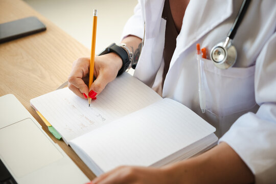 A medical professional in a white coat writes notes in a journal with a pencil, a stethoscope around their neck, and a laptop nearby.