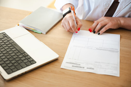 A person in a white coat is writing on a document at a desk with a laptop and a notebook.