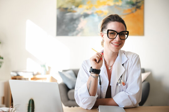A smiling female doctor in a white coat sits at her desk with a laptop and holds a pencil, ready to assist patients.