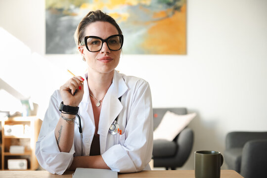 A female doctor in a white coat sits at a desk, holding a pencil and looking at the camera.