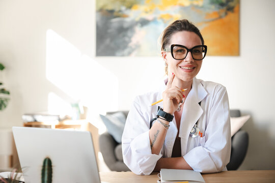 A smiling female doctor in a white coat sits at her desk with a laptop and notebook, holding a pencil to her chin.