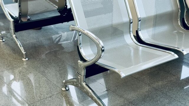 Empty metal benches reflecting polished floor, lone passenger seated cross-legged using phone, beige outfit, black bag at feet