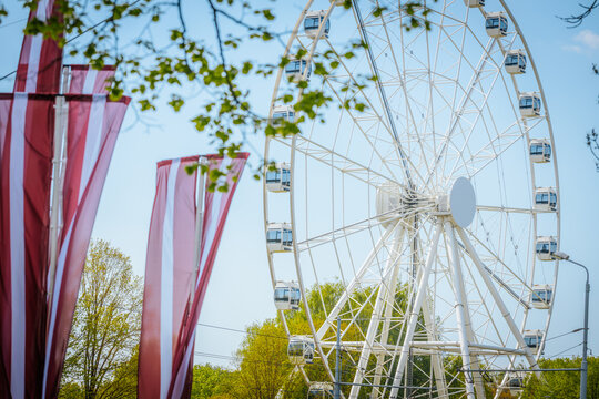 Large white Ferris wheel with blue sky backdrop and colorful flags in foreground, located in a public park during a festive outdoor event in Latvia