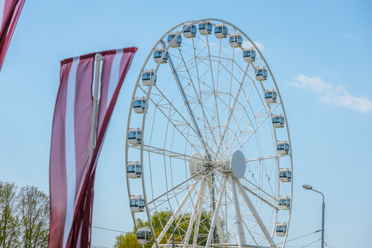 Large white Ferris wheel with blue cabins against a clear sky, colorful flags waving in the foreground, trees and streetlight visible in the background