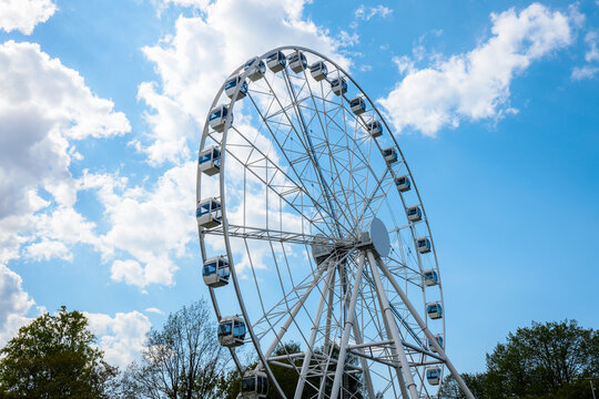 Large white Ferris wheel against a bright blue sky with fluffy clouds, surrounded by green trees in a public amusement park setting