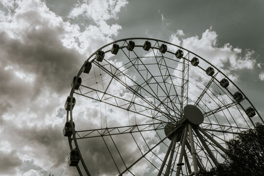 Large Ferris wheel silhouette against a cloudy sky, showcasing intricate metal framework and passenger cabins, surrounded by trees in an outdoor amusement park setting