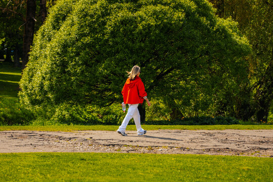 Woman in red jacket walks along a path in a park with lush green trees and grass, enjoying a sunny day outdoors in a natural setting