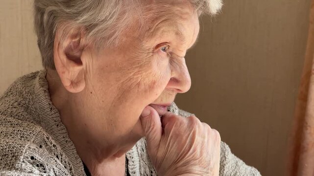 An elderly grandmother dressed in a knitted sweater is sitting in a room, thoughtfully looking away, holding her hand to her chin. Planning of actions. Close-up