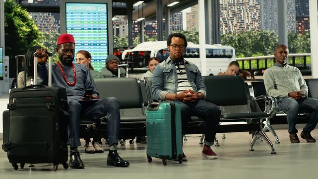 African American solo traveler in bus terminal surrounded by passengers, having trolley bag and backpack. Man checks departure time information on screen before boarding interstate bus.