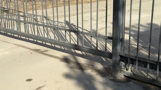 Close-up iron gate motor, spring day. Parking entrance automation in sunlight, metal bars casting elongated shadows on concrete
